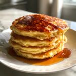 A stack of fluffy buttermilk pancakes drizzled with golden syrup on a white plate.
