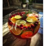 A large glass bowl filled with festive Christmas Punch, featuring slices of oranges, limes, fresh cranberries, and a sprig of rosemary.
