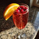 A tall glass filled with a bubbly Cranberry Mimosa, topped with cranberries and garnished with an orange slice.