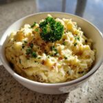 A bowl of creamy mashed potatoes with roasted garlic, garnished with parsley and crispy bits.