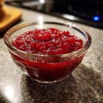 A close-up of a small glass bowl filled with vibrant, homemade Cranberry Sauce.