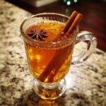 A close-up of a warm Hot Toddy in a glass mug, garnished with cinnamon sticks and star anise.