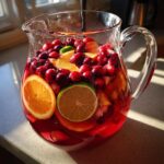 A glass pitcher filled with Sparkling Cranberry Punch, garnished with fresh cranberries, orange slices, and lime slices.