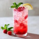 A refreshing Sparkling Raspberry Lemonade Mocktail in a tall glass, filled with ice, raspberries, mint, and a lemon slice.