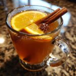 A clear mug filled with warm Spiced Apple Cider, garnished with orange slices, cinnamon sticks, and star anise.