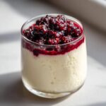 A close-up of creamy Vanilla Bean Panna Cotta topped with a vibrant berry compote in a glass.