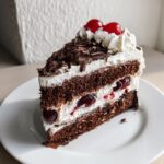 A slice of Black Forest Cake on a white plate, showing layers of chocolate cake, whipped cream, and cherries.