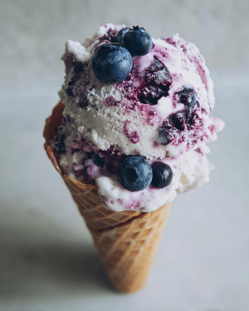 Close-up of a scoop of blueberry yogurt ice cream in a waffle cone, topped with fresh blueberries.
