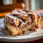 A close-up of a slice of Cinnamon Roll Coffee Cake with icing on a white plate.