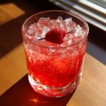 A vibrant red love potion cocktail in a glass, garnished with a fresh raspberry on a skewer, served with ice.