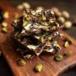 Close-up of stacked White Chocolate Pistachio Bark pieces on a wooden board, with pistachios scattered around.