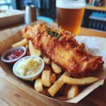 A plate of delicious Beer-Battered Fish and Chips with tartar sauce, ketchup, lemon, and a pint of beer.