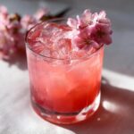 A vibrant Cherry Blossom Mocktail in a glass, garnished with pink cherry blossoms and ice.