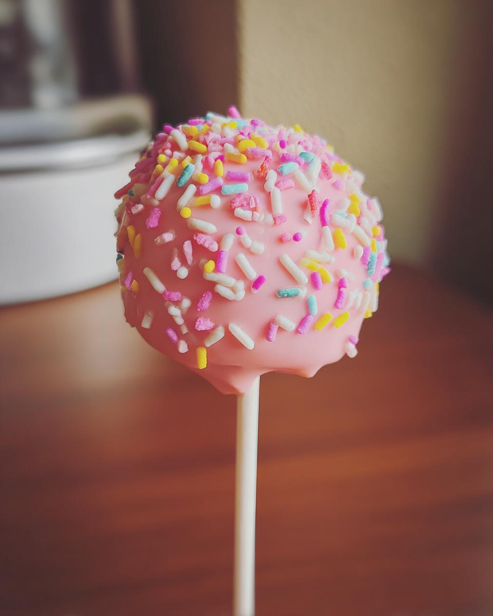 Close-up of a pink Donut Hole Cake Pop covered in colorful sprinkles on a stick.