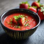 Close-up of a bowl of Strawberry Basil Gazpacho, garnished with a strawberry slice and herbs.