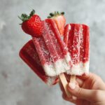 Hand holding several Strawberry Coconut Dog Popsicles, decorated with fresh strawberries and coconut flakes.