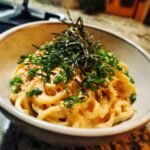 A bowl of creamy Mentaiko Udon, a Japanese noodle dish with salty fish roe, garnished with nori and scallions.