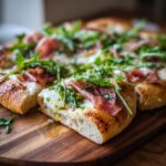 Close-up of a slice of Prosciutto and Pea Shoot Pizza on a wooden board, showing toppings.