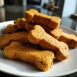 Close-up of a pile of bone-shaped Gluten-Free Pumpkin Dog Biscuits on a white plate.