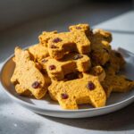 Pile of homemade Pumpkin Cranberry Holiday Dog Biscuits on a white plate, shaped like dogs.