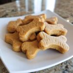 Pile of bone-shaped Pumpkin Peanut Butter Cut-Out Dog Biscuits on a white plate.